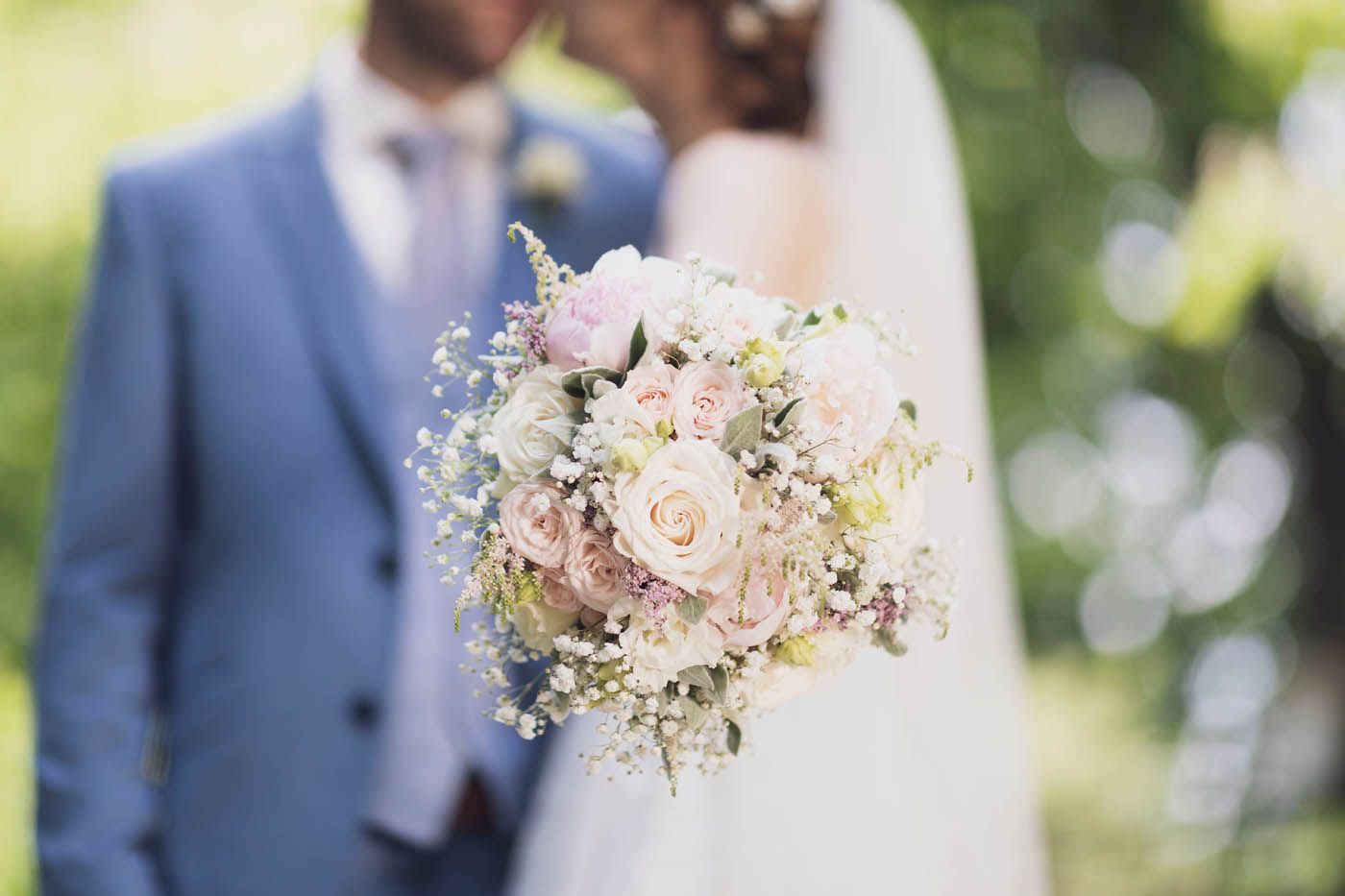 Brautstrauß im Fokus; Paar im Hintergrund verschwommen, Bräutigam im blauen Anzug, Braut mit Schleier.
