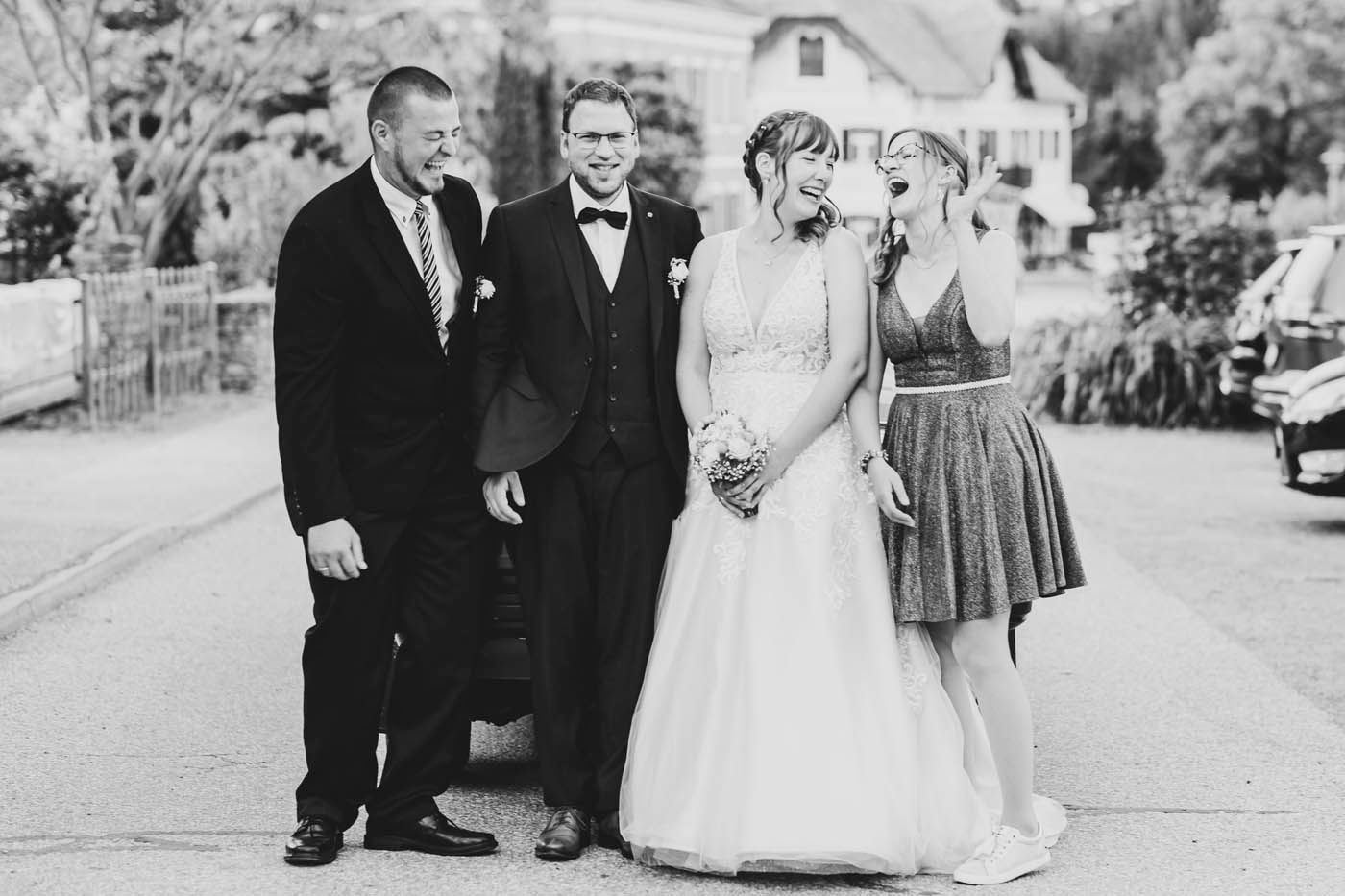 Hochzeitsgesellschaft lacht auf der Straße. Braut im Kleid, Bräutigam im Anzug.