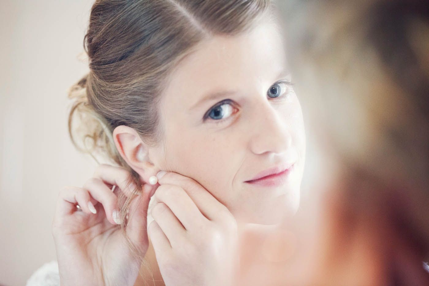 Frau legt Perlenohrringe an, Haare hochgesteckt. Sanftes Licht, neutraler Hintergrund, Blick in den Spiegel.