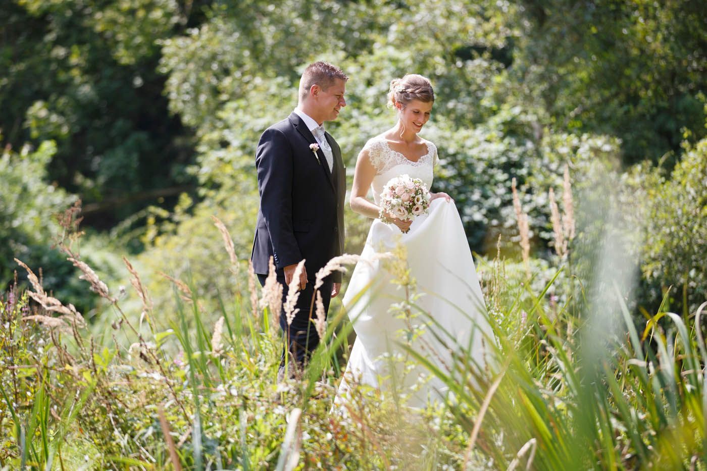Braut und Bräutigam schreiten durch hohes Gras in sonniger Umgebung. Die Braut trägt ein weißes Kleid