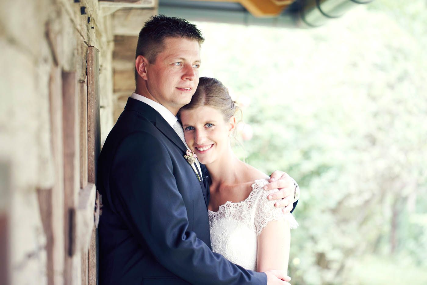 Ein Paar in Hochzeitskleidung umarmt sich lächelnd und lehnt im Freien an einer holzgetäfelten Wand.