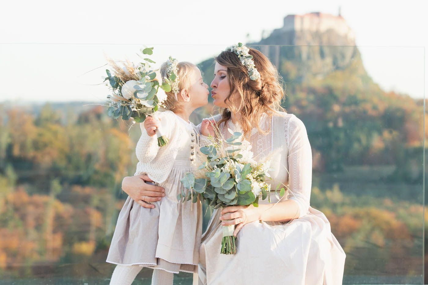 Eine Frau in einem weißen Kleid küsst ein junges Mädchen. Beide halten Blumensträuße und tragen Blumenkränze