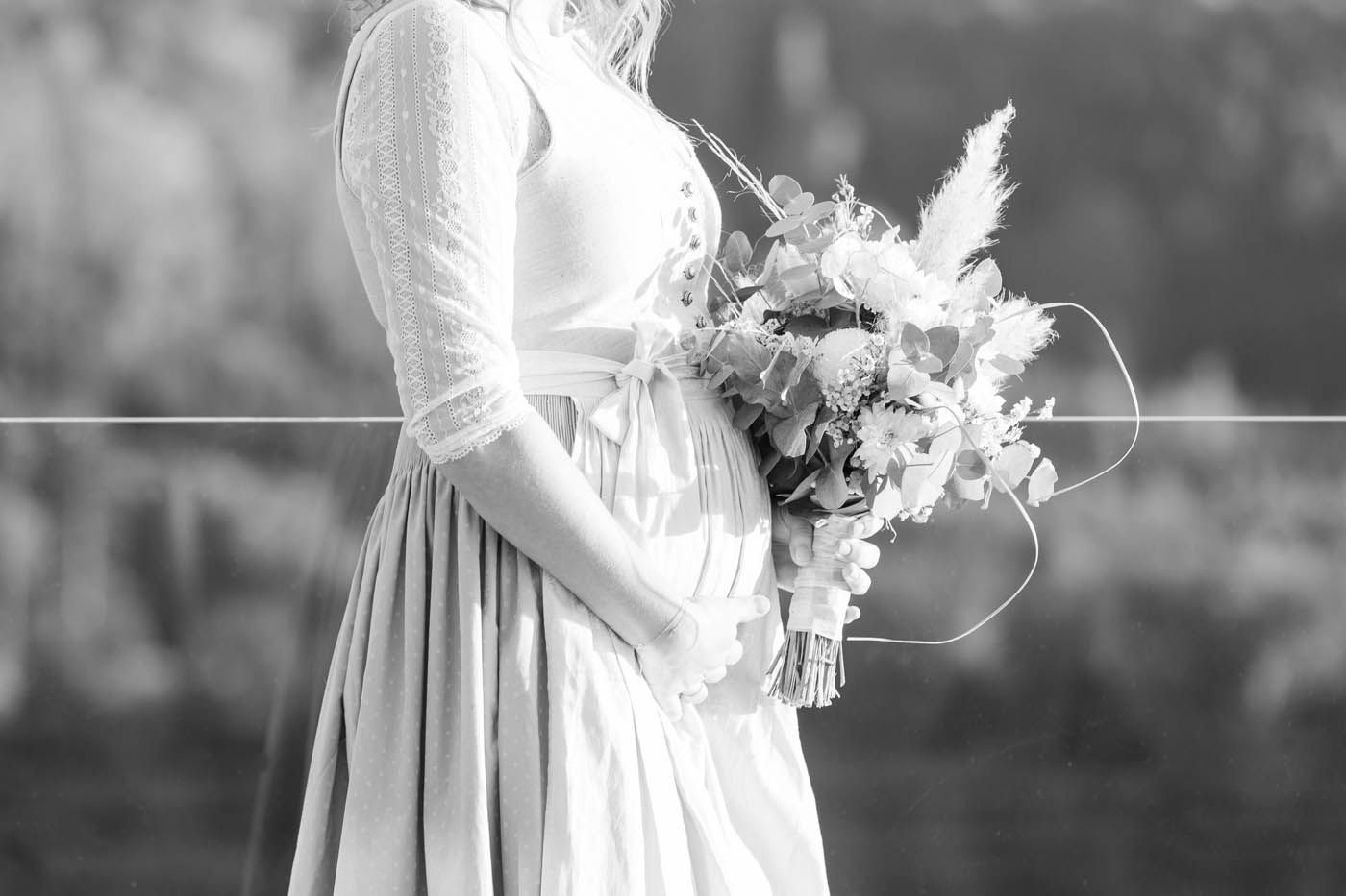 Frau mit Blumenstrauß, bekleidet mit einem weißen Kleid und Bolero, die Hände auf dem Bauch, im Freien.