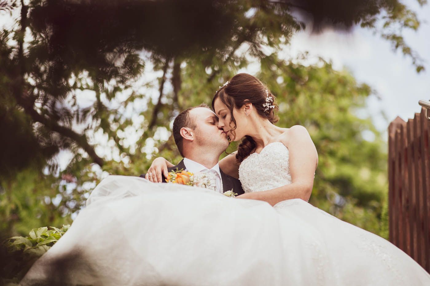 Braut und Bräutigam küssen sich im Freien; sie trägt ein weißes Kleid, er einen Anzug. Grünes Laub umrahmt das Paar.