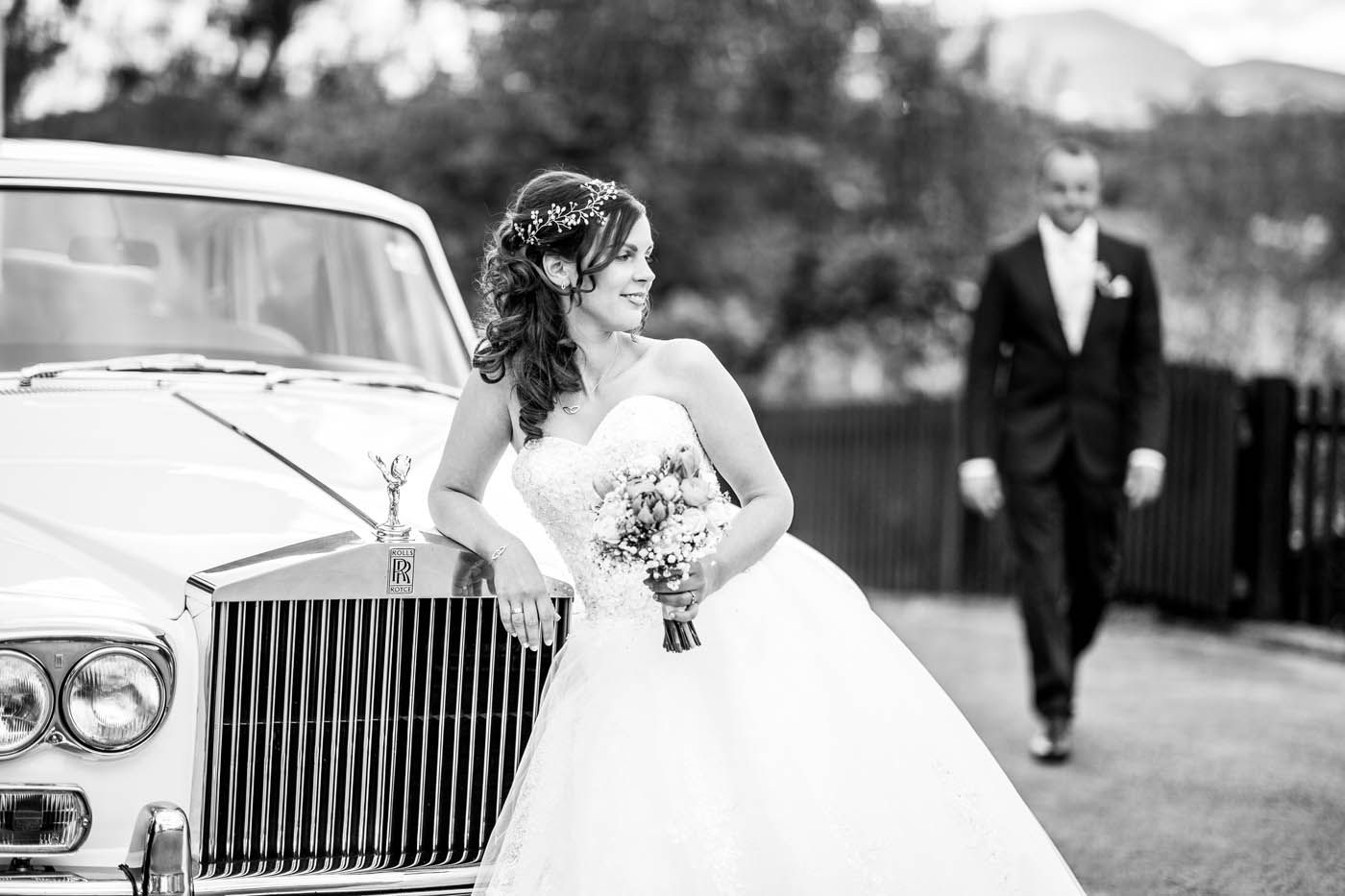 Die Braut im weißen Kleid lehnt an einem Oldtimer; der Bräutigam im schwarzen Anzug nähert sich.