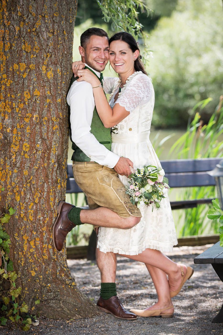 Ein Paar in traditioneller deutscher Hochzeitskleidung lächelt und umarmt sich in der Nähe eines Baumes und eines Gewässers.