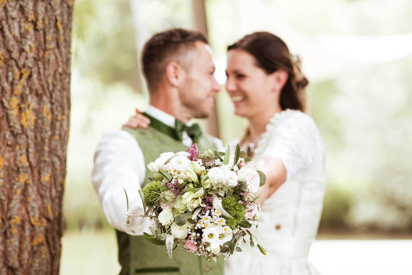 Brautpaar in traditioneller Kleidung, das einen Brautstrauß hält und im Freien lächelt.