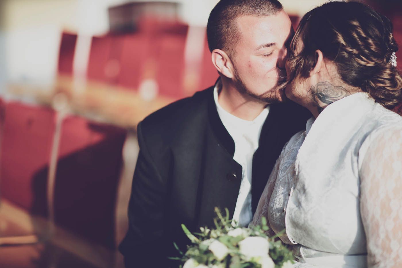 Küssendes Paar, Braut hält Blumen, Bräutigam im Anzug, verschwommener roter Hintergrund.