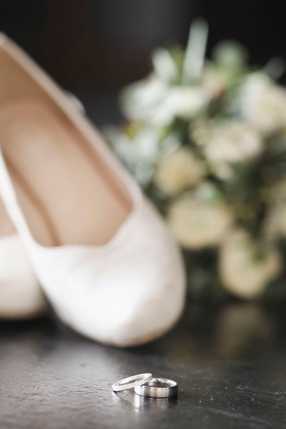 Im Fokus die Eheringe, im Hintergrund sind Brautschuhe und Brautstrauß verschwommen zu sehen.
