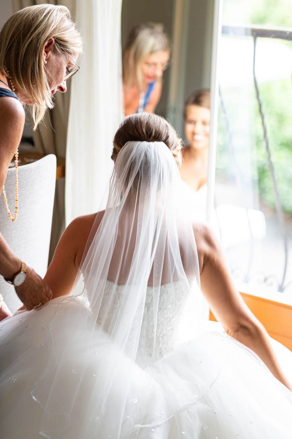 Die Braut im Hochzeitskleid und Schleier wird von einer Frau unterstützt; zwei Frauen schauen vom Fenster aus zu.