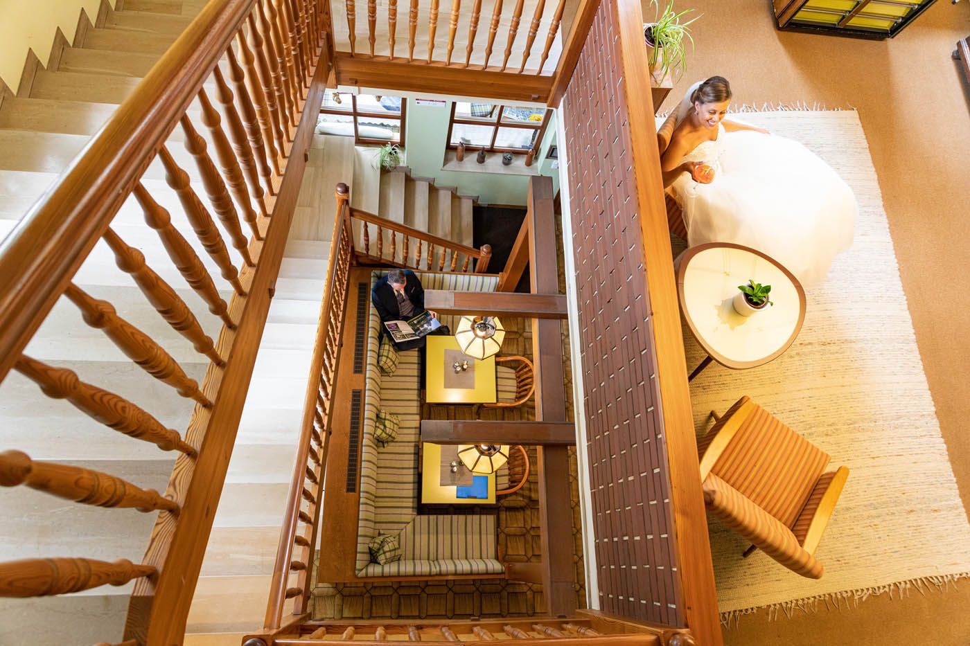 Draufsicht auf ein Treppenhaus mit hellem Holzgeländer. Eine Frau sitzt auf einem Sofa neben einem runden Tisch.