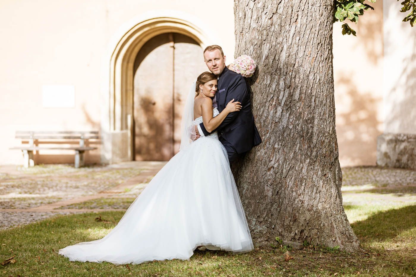 Ein Brautpaar umarmt sich neben einem Baum, im Hintergrund ist ein Gebäude mit einem Bogeneingang zu sehen.