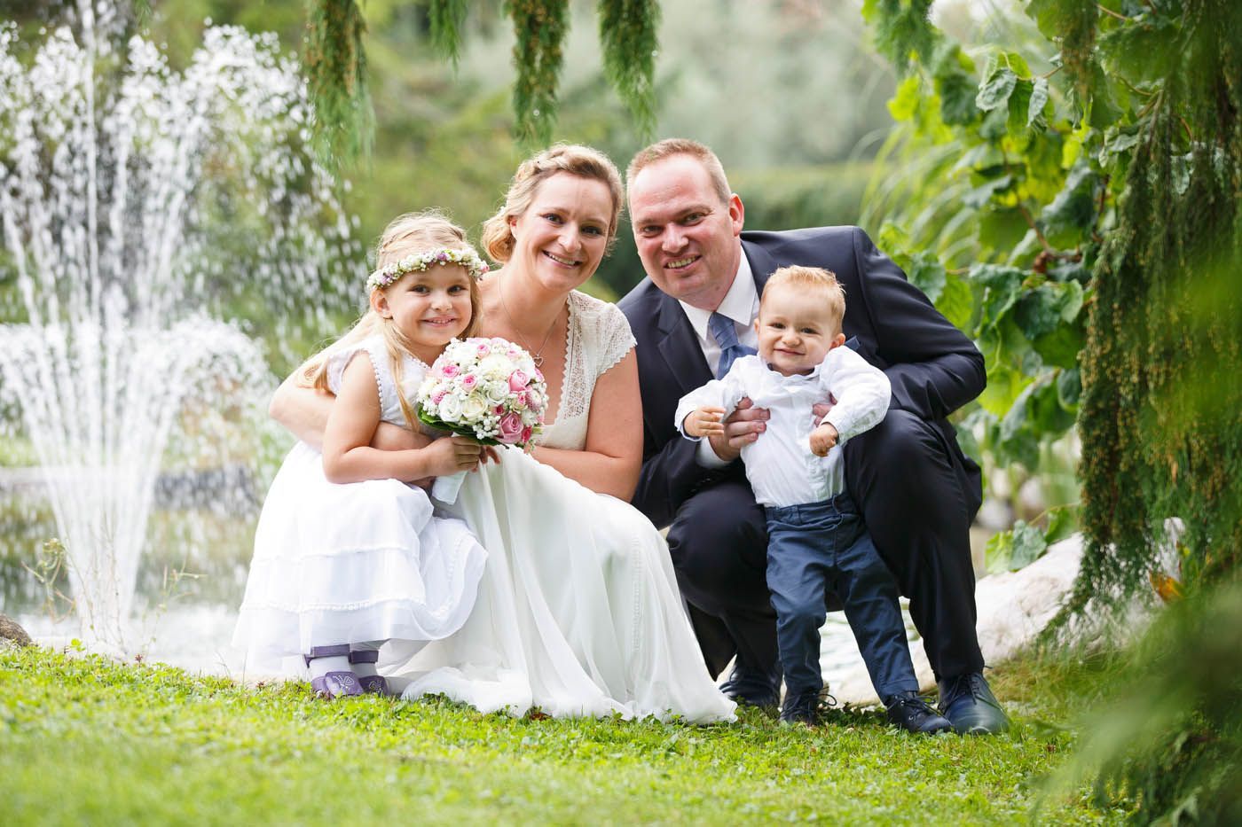 Familienporträt mit Braut, Bräutigam und zwei Kindern, die in der Nähe eines Brunnens und im Grünen posieren