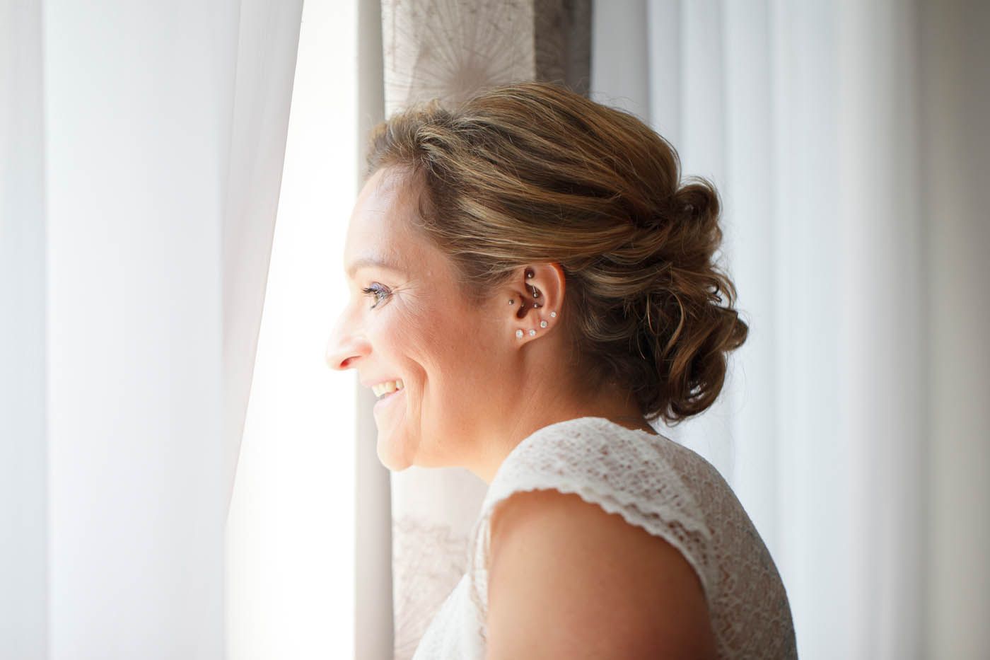 Frau mit hochgesteckter Frisur lächelt in der Nähe eines Fensters; sie trägt ein weißes, spitzenbesetztes Oberteil.