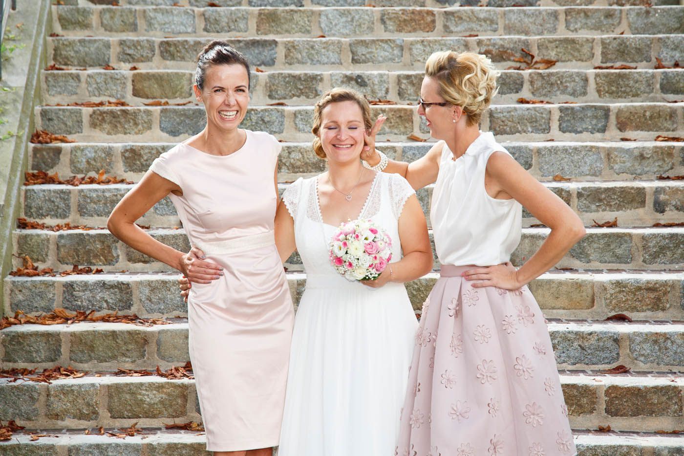 Braut und Brautjungfern auf Steinstufen; Braut hält den Brautstrauß, Brautjungfern lächeln.
