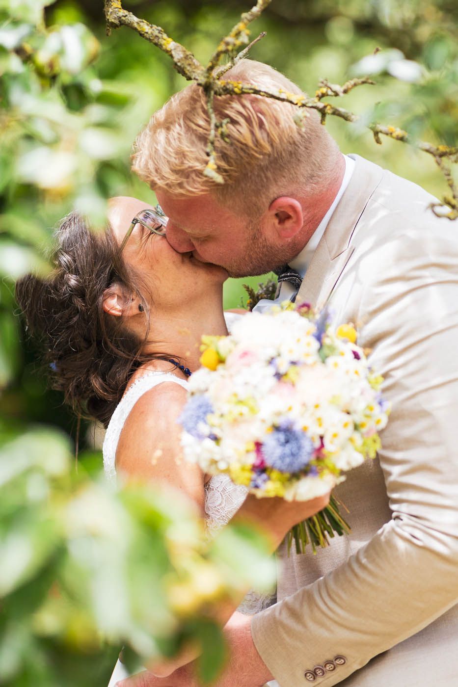 Ein Paar küsst sich, sie hält einen Blumenstrauß, umrahmt von Ästen und Laub.