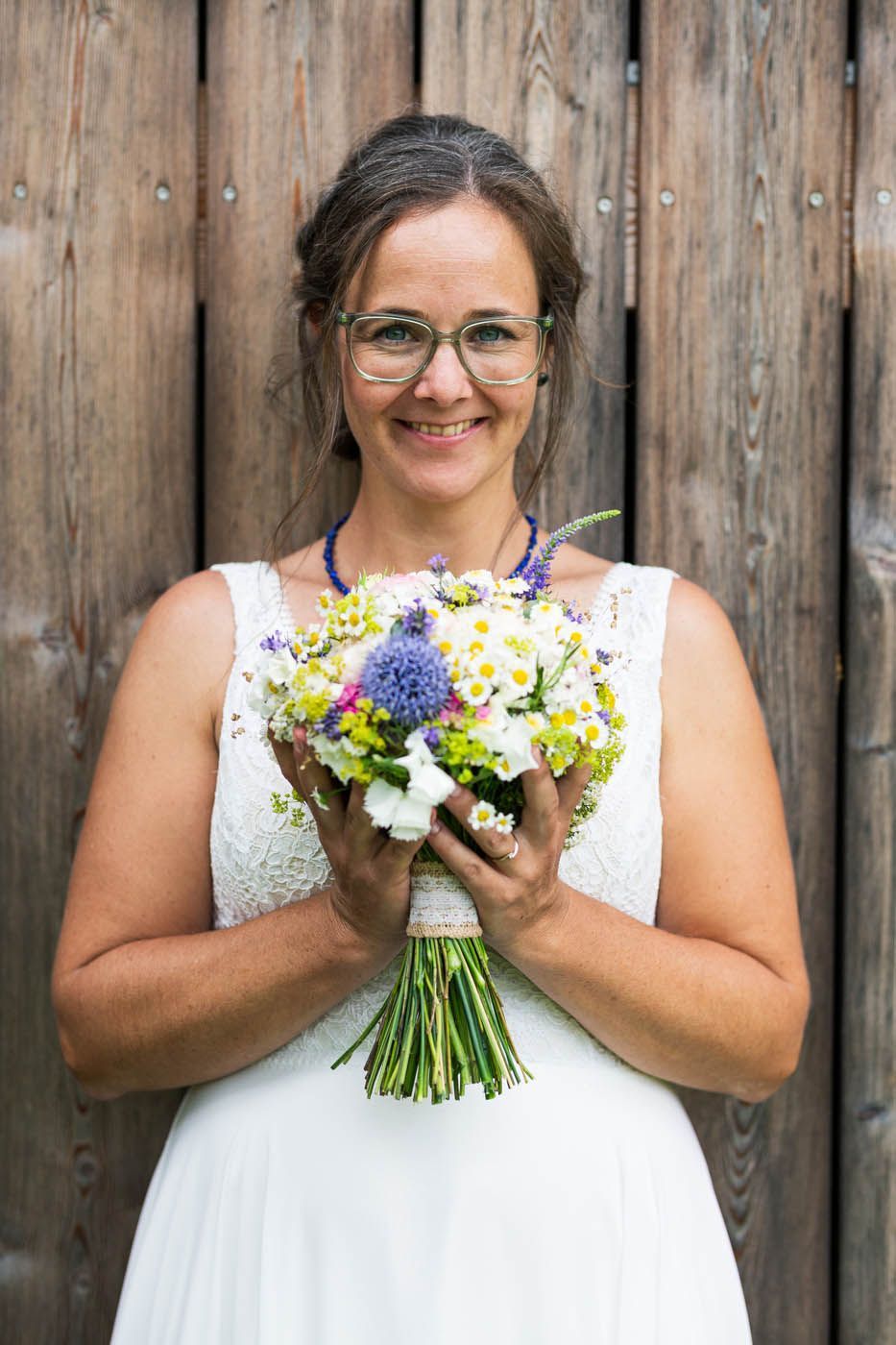 Frau in einem weißen Kleid, die einen Blumenstrauß hält, lächelt vor einer Holzwand.