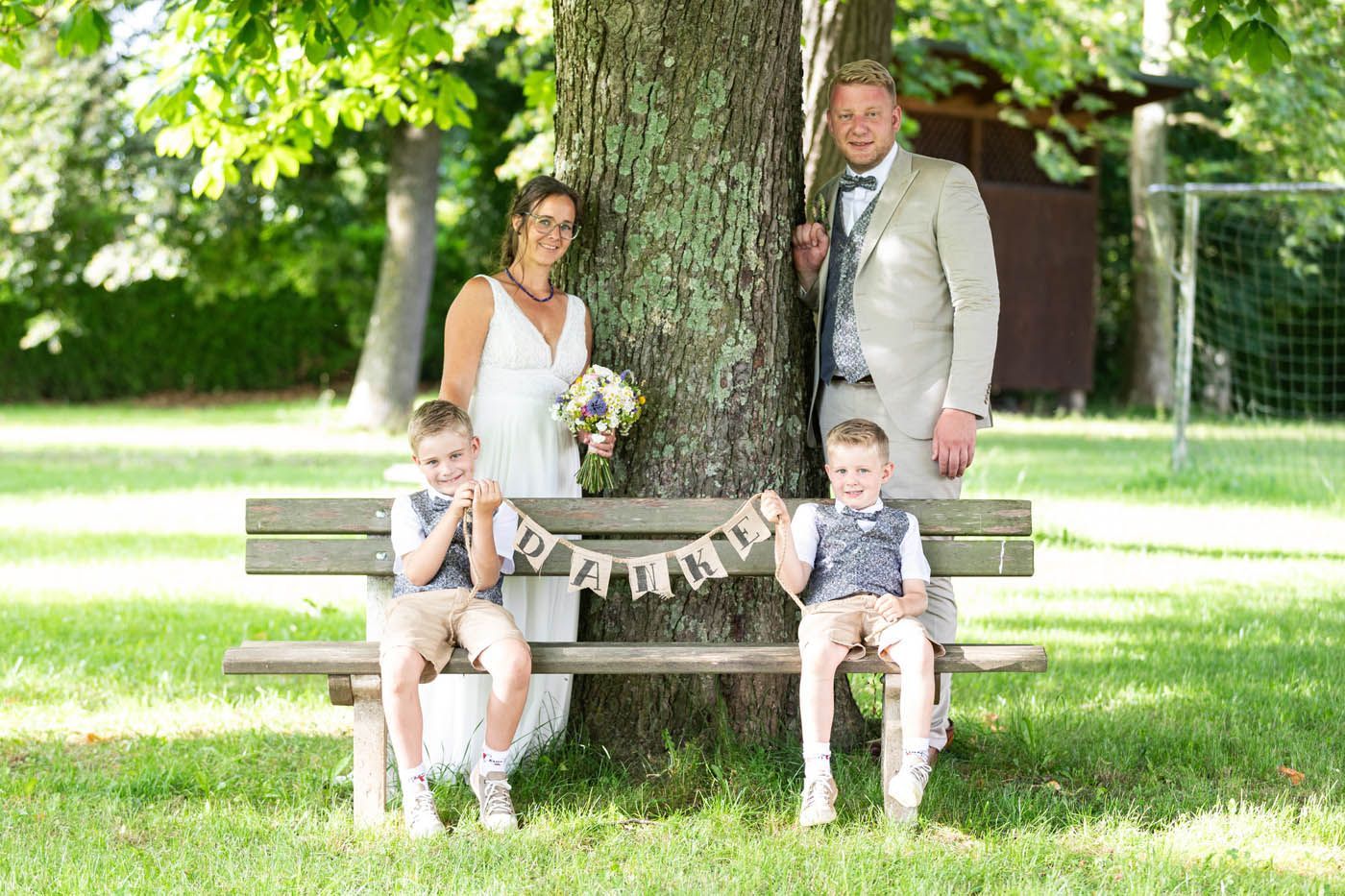 Hochzeitsfoto: Paar mit zwei Jungen auf einer Parkbank. Auf dem Banner steht „JA“.