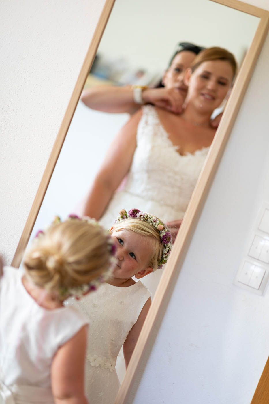 Braut und Blumenmädchen im Spiegel. Frau im Brautkleid mit Begleiterin, kleines Mädchen mit Blumenkranz lächelt.