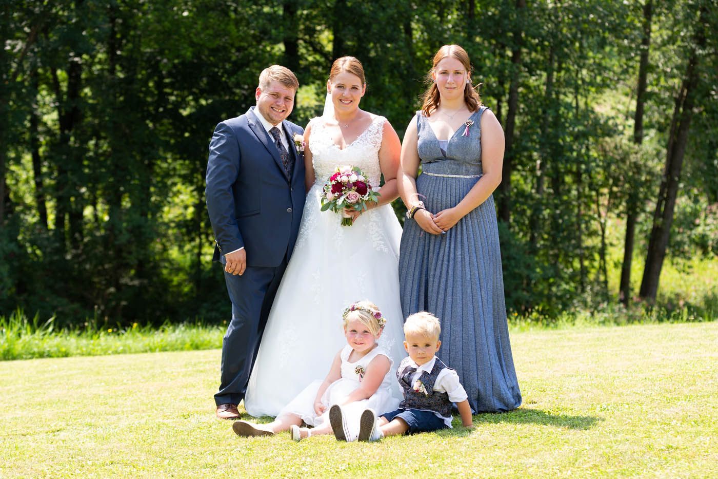 Hochzeitsgruppe posiert im Freien; Brautpaar, Brautjungfer und zwei Kinder. Grüne Umgebung.
