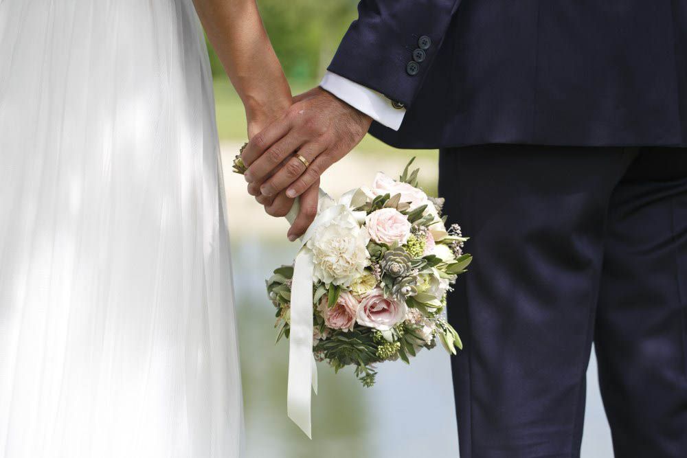 Brautpaar hält Händchen, trägt Brautstrauß, in der Nähe eines Gewässers.