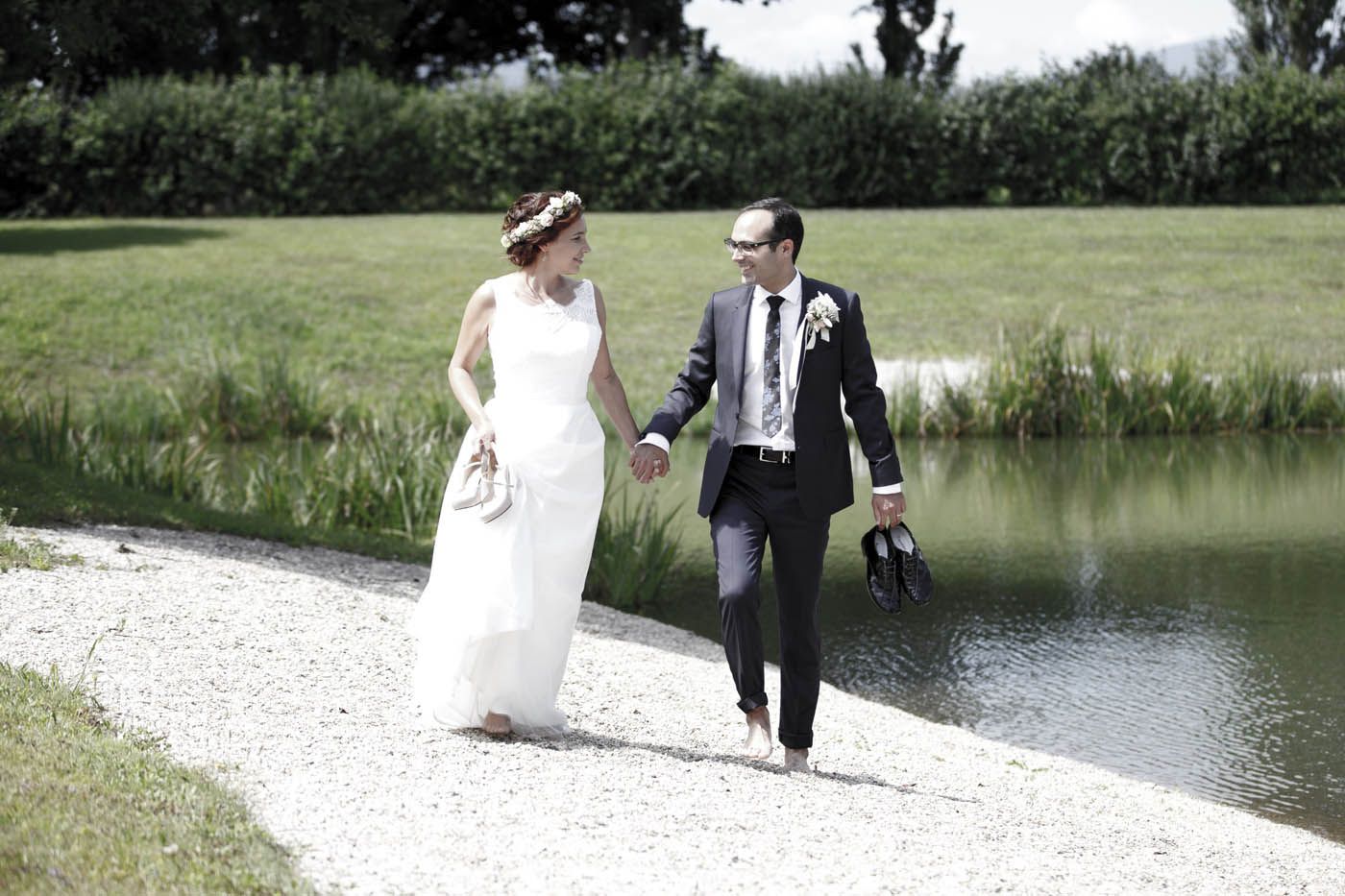Braut und Bräutigam spazieren an ihrem Hochzeitstag barfuß und Hand in Hand an einem See entlang.