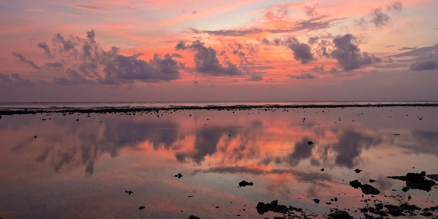 Sonnenuntergang über ruhigem Wasser, in dem sich der Himmel spiegelt, mit Wolken in Rosa-, Orange- und Violetttönen.
