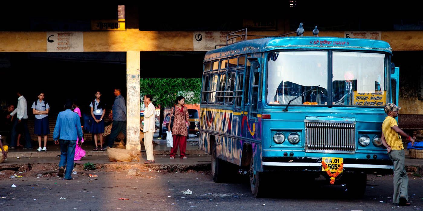 Ein blauer Bus steht an einer Bushaltestelle, Menschen warten unter einem Unterstand. Ein Mann lehnt an dem Bus.