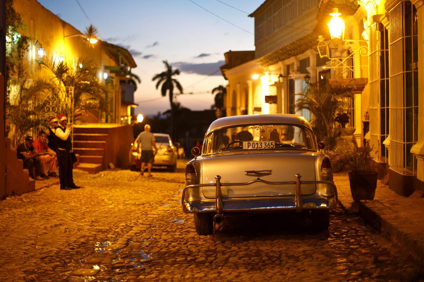 Ein Oldtimer parkt in der Abenddämmerung auf einer Kopfsteinpflasterstraße in Kuba, beleuchtet von Straßenlaternen.