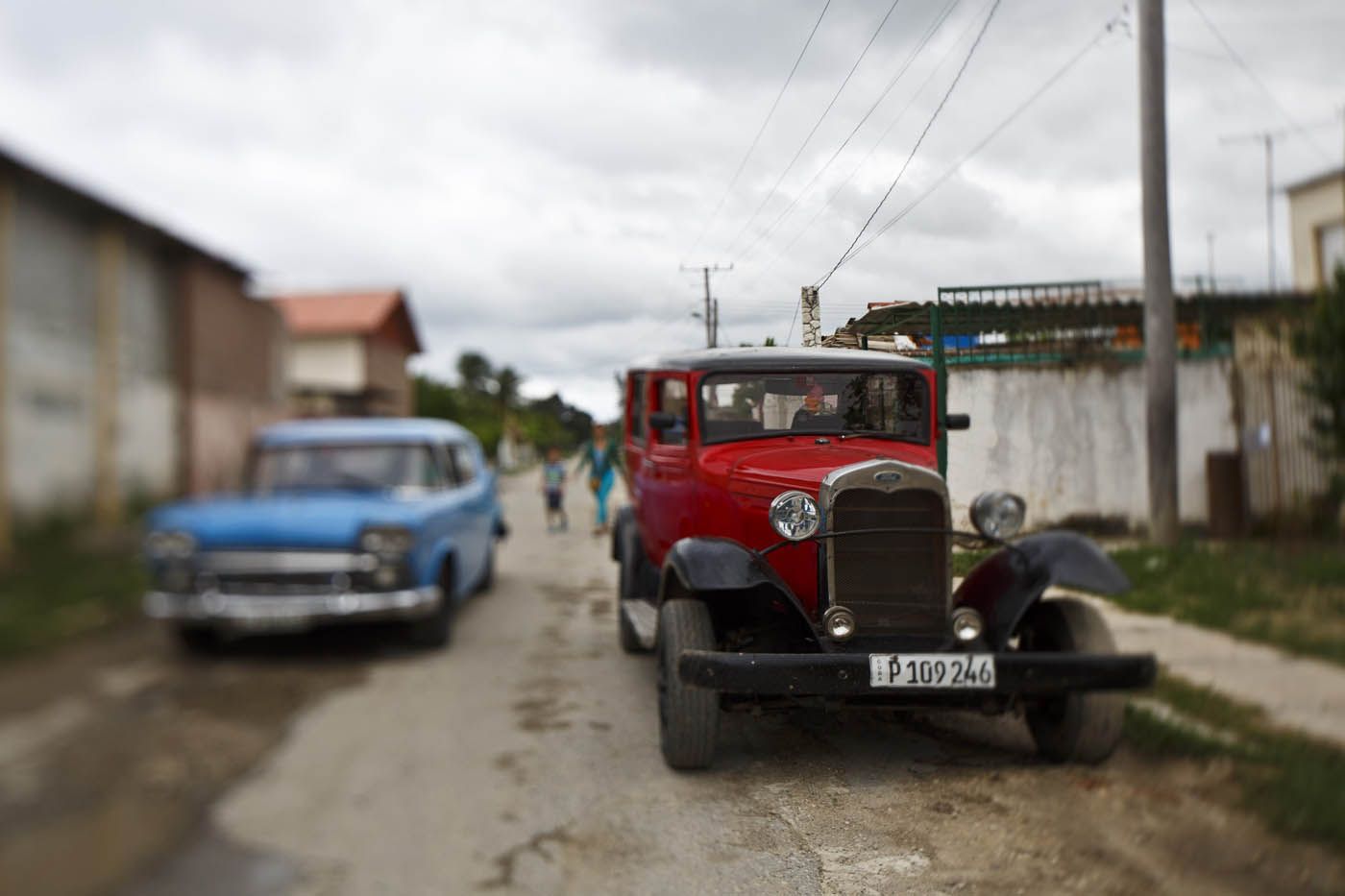 Ein roter Oldtimer auf einer unbefestigten Straße in Kuba, im Hintergrund ein blaues Auto und verschwommene Gestalten.