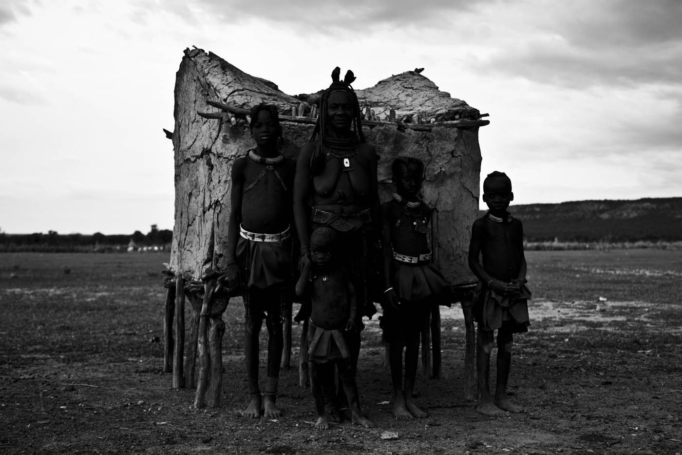 Eine Familie steht vor einer strohgedeckten Hütte in einer ländlichen, trockenen Umgebung. Schwarz-Weiß-Fotografie.
