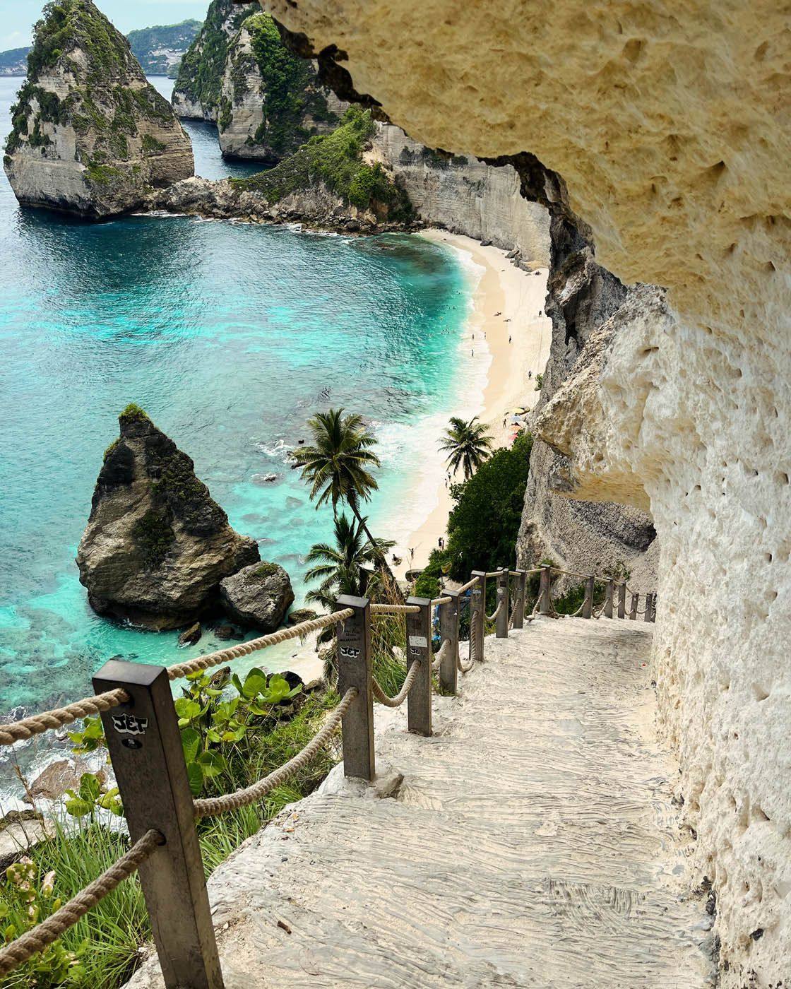 Treppe, die zu einem weißen Sandstrand mit türkisfarbenem Wasser, Klippen und Felsformationen in tropischer Umgebung führt.