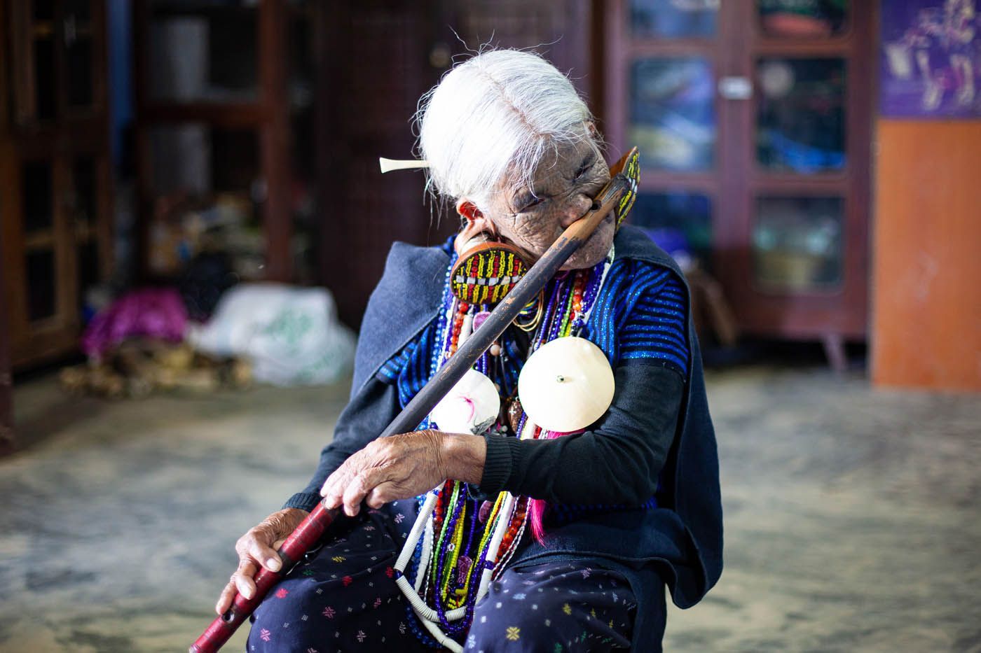 Eine ältere Person spielt drinnen eine lange Holzflöte. Sie trägt bunte Perlen und ein dunkles Oberteil.