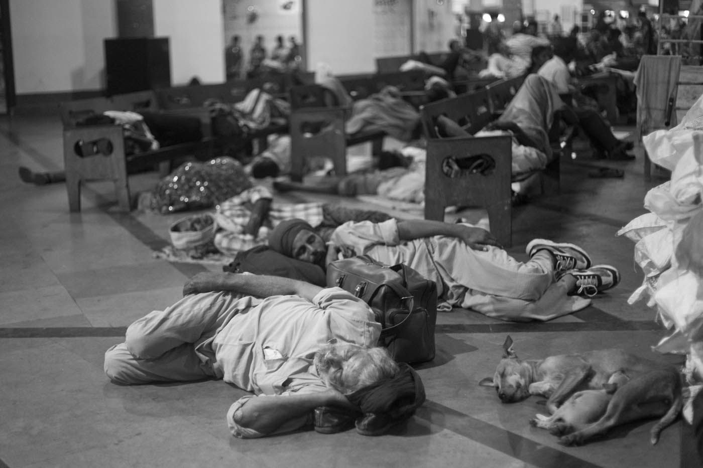 Menschen schlafen auf dem gefliesten Boden, einige haben ihre Habseligkeiten in der Nähe. Ein paar Stühle sind zu sehen.