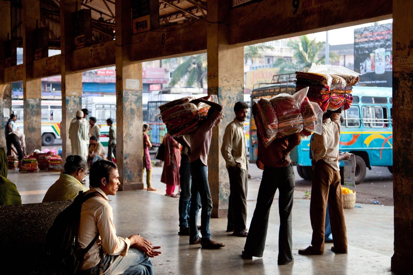 Menschen mit Paketen auf dem Kopf an einem Busbahnhof. Im Hintergrund ein blauer Bus.
