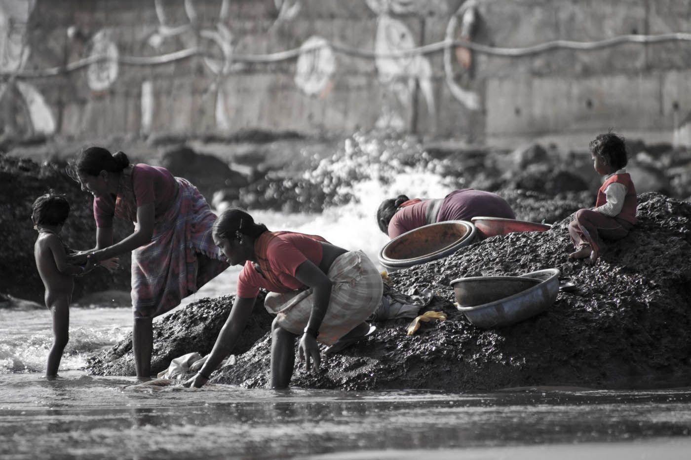 Frauen und ein Kind waschen Wäsche in einem Fluss, daneben stehen Schüsseln. Im Hintergrund eine Betonmauer.
