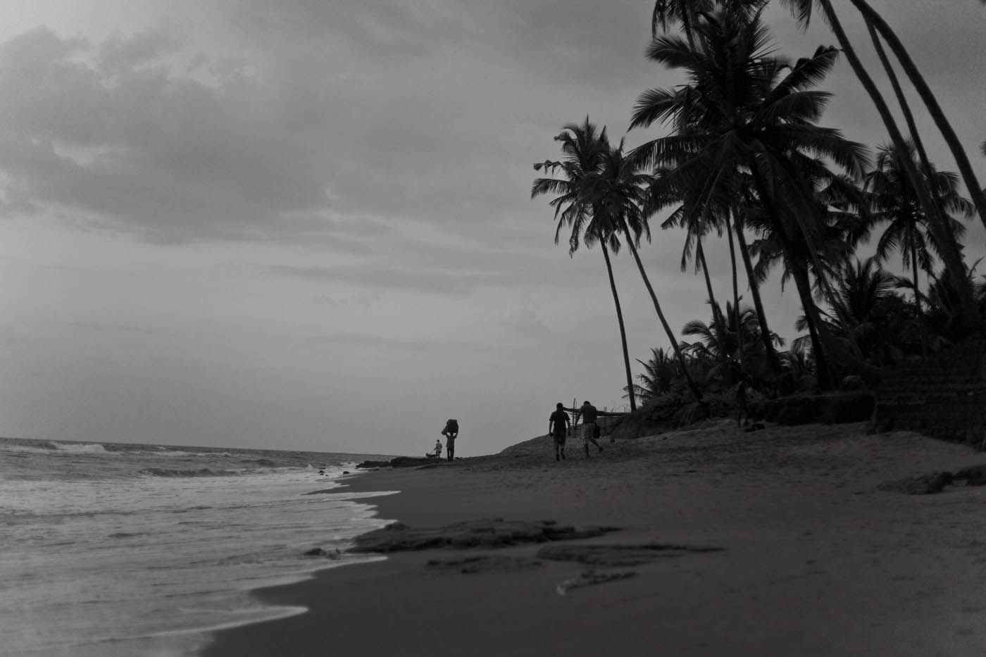 Strandszene in Schwarz-Weiß. Palmen säumen das Ufer, Wellen brechen sich am Strand, Menschen spazieren am Ufer entlang. Bewölkter Himmel.
