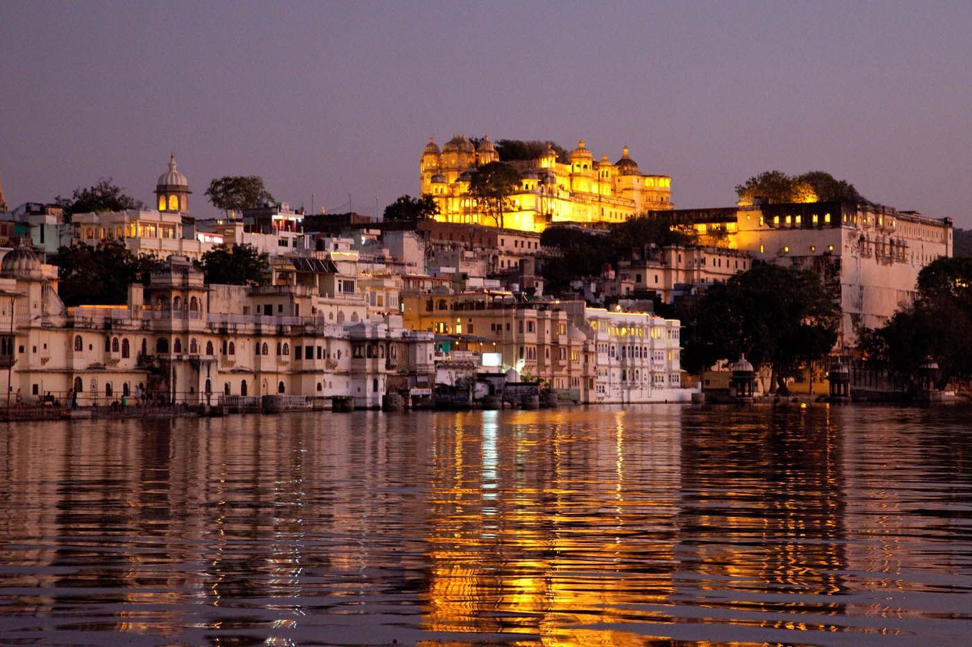Nachtansicht von beleuchteten weißen Gebäuden am Ufer eines Sees, goldene Lichter spiegeln sich im Wasser.
