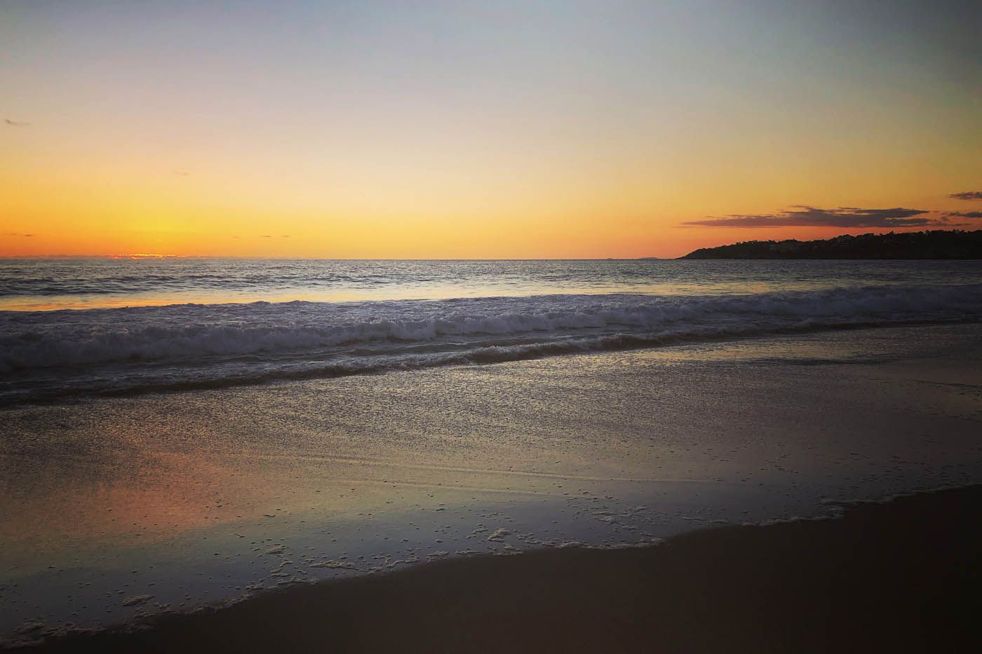 Sonnenuntergang über den Wellen des Ozeans, der warme orange und gelbe Himmel spiegelt sich im nassen Sand.
