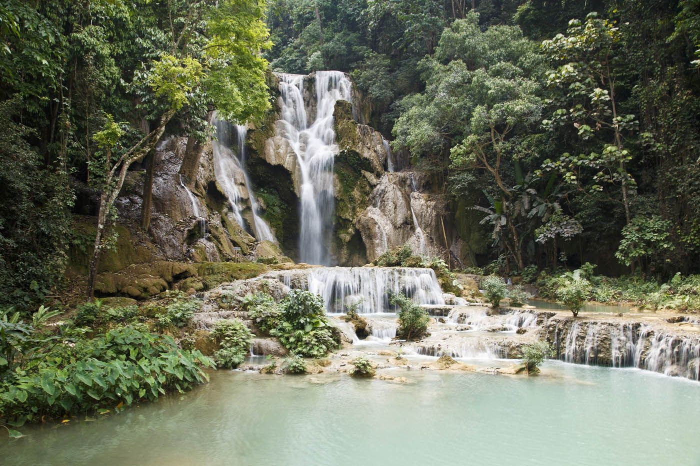 Ein Wasserfall stürzt über terrassenförmige Felsformationen in ein türkisfarbenes Becken, umgeben von üppigem Grün.