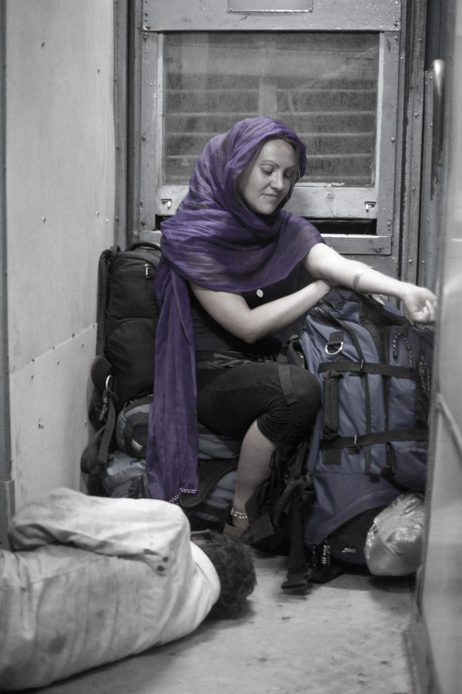 Eine Frau im Zug, umgeben von Gepäck, trägt einen lila Schal. Sie sitzt eng im Abteil und lächelt.