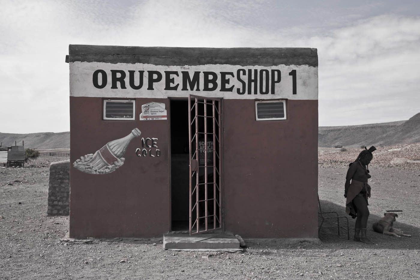 Kleiner Laden in einer Wüstenlandschaft mit einem Flaschenmotiv und einer daneben stehenden Person.