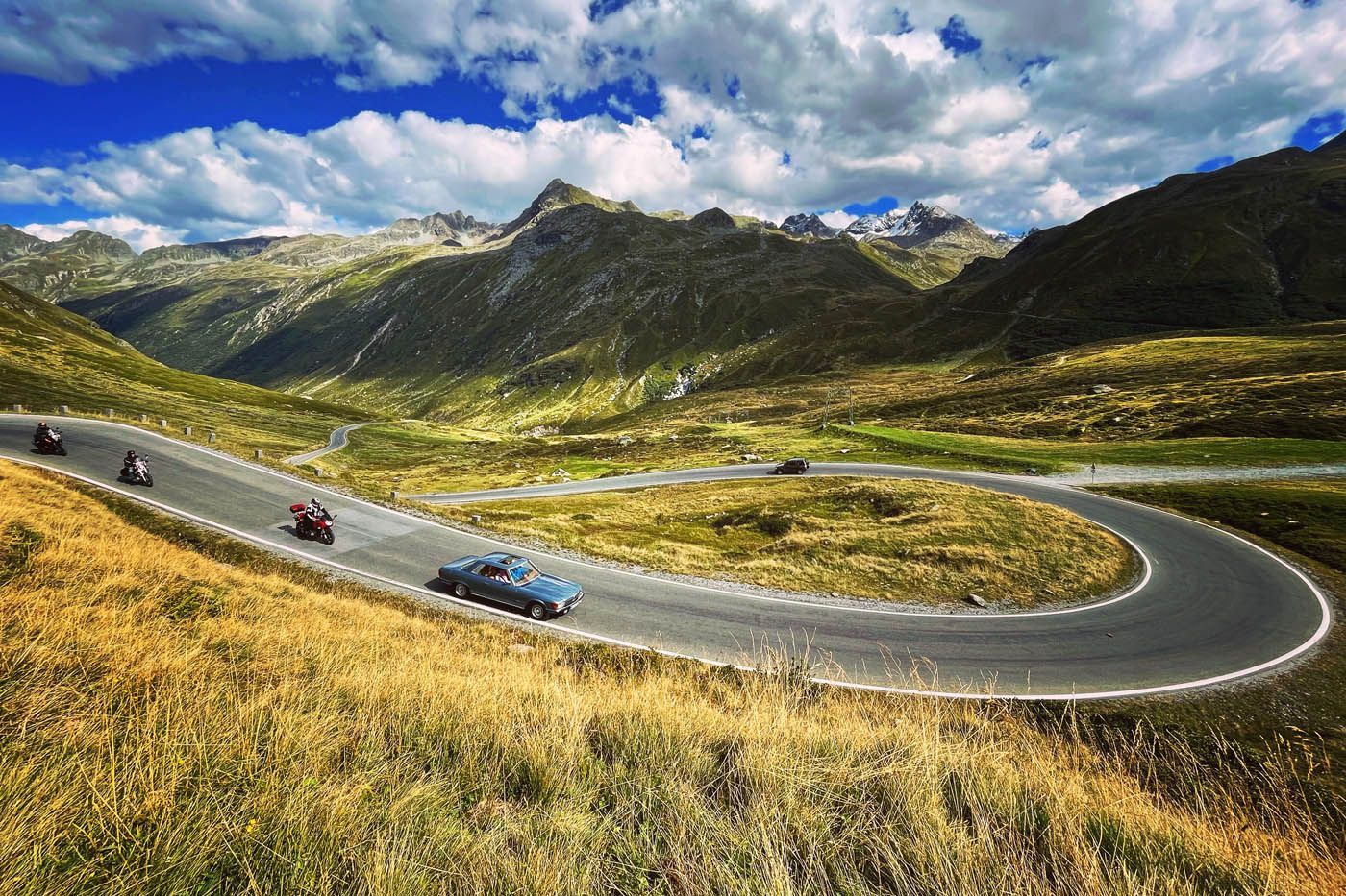 Kurvenreiche Bergstraße mit einem blauen Auto und Motorrädern. Üppige grüne und braune Landschaft unter blauem Himmel.