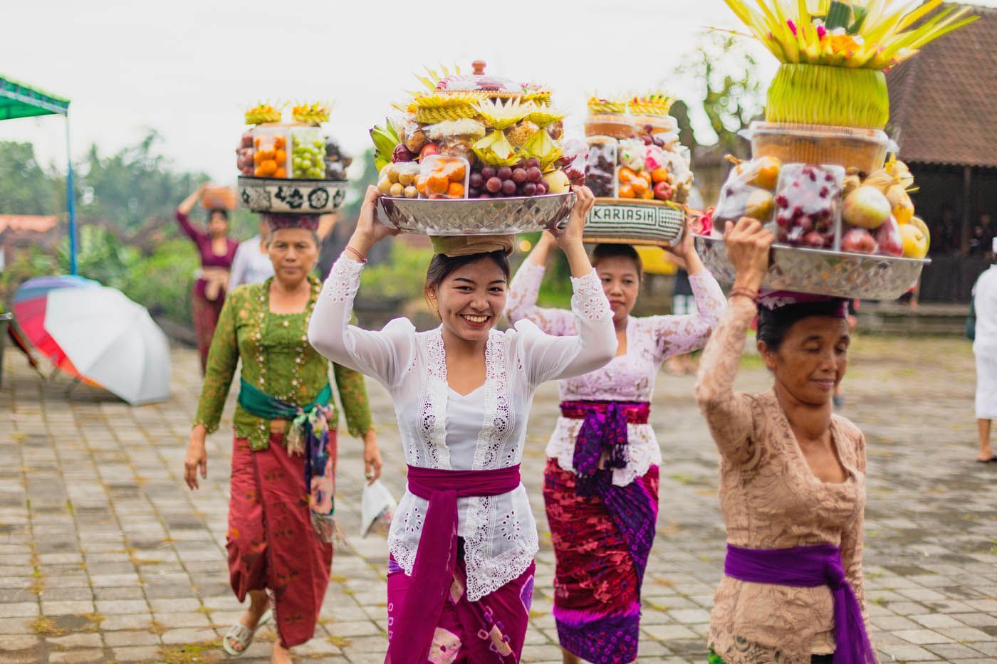 Frauen in traditioneller Tracht tragen lächelnd Fruchtopfer auf dem Kopf. Im Hintergrund ein Tempel.