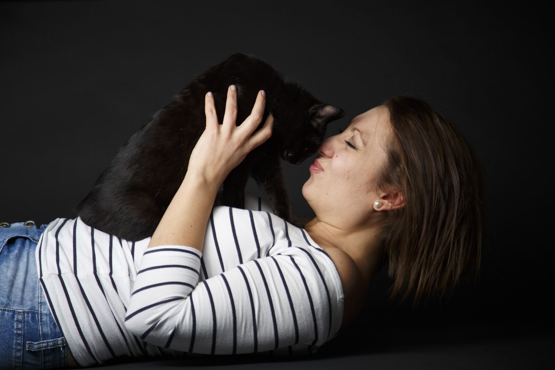 Frau liegt vor dunklem Hintergrund und küsst eine schwarze Katze.