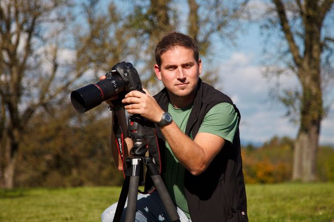 Ein Mann, der im Freien eine Kamera auf einem Stativ justiert, trägt ein grünes Hemd und eine dunkle Weste.