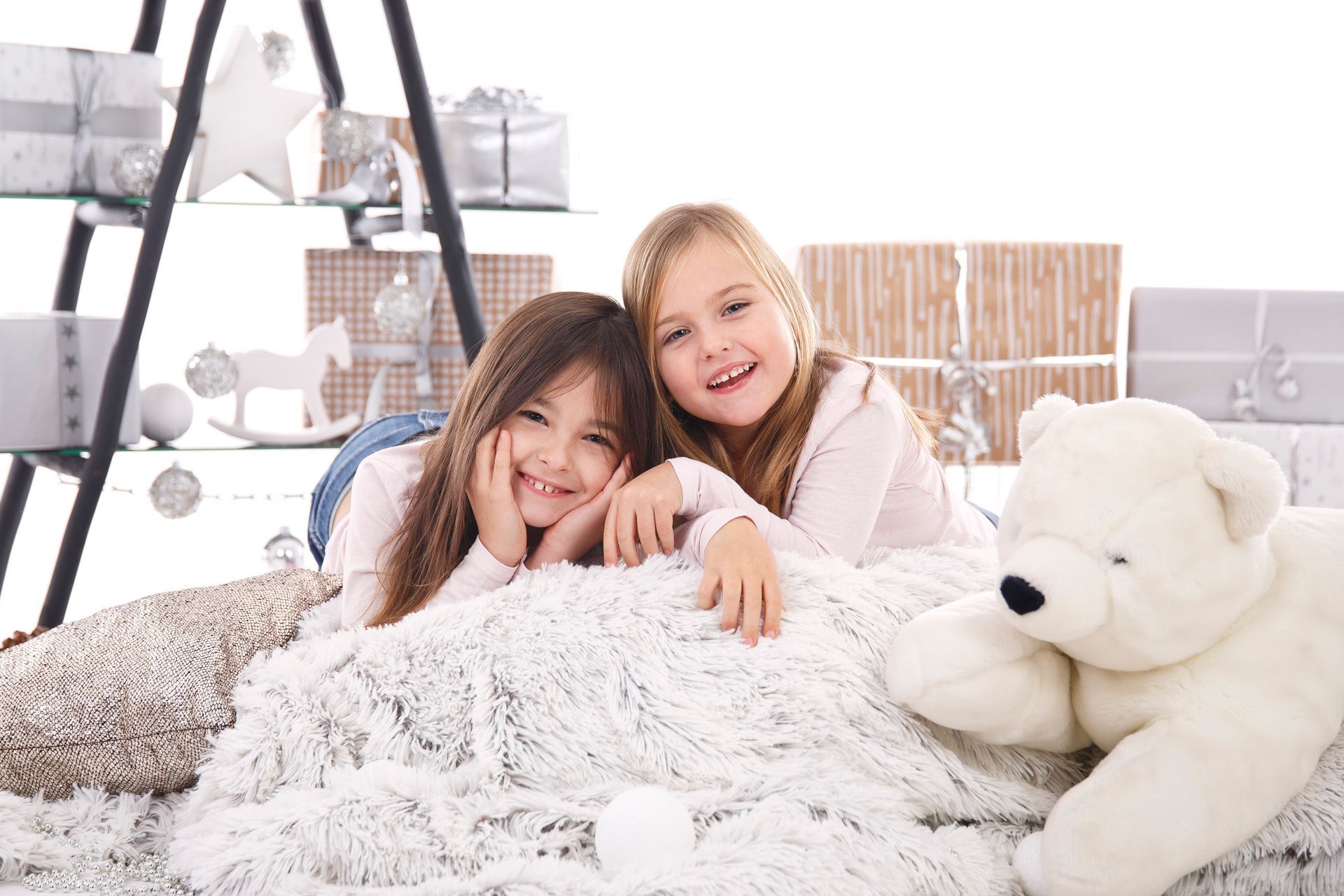 Zwei lächelnde Mädchen liegen auf einer flauschigen Decke neben einem Teddybären und Geschenken.