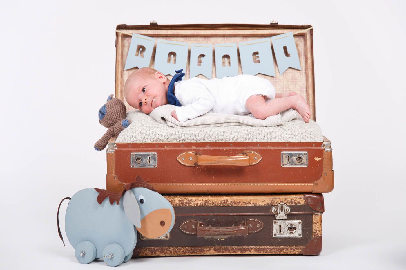 Baby liegt auf Koffern mit Stofftieren, darüber ein hellblaues Banner mit der Aufschrift „Rachael“.