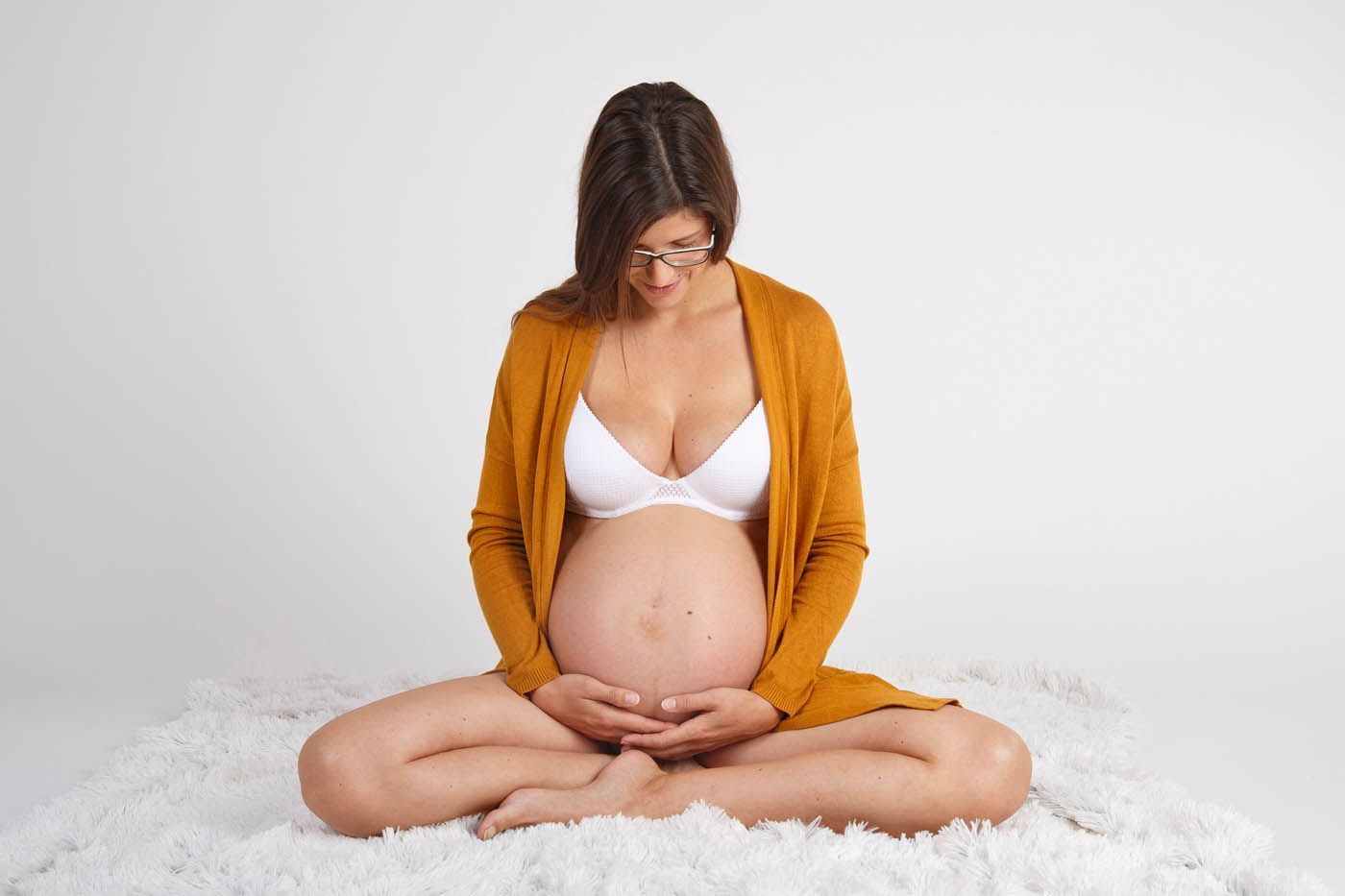 Schwangere Person, Hände auf dem Bauch, trägt BH und offene Strickjacke, sitzt auf einer flauschigen weißen Oberfläche.