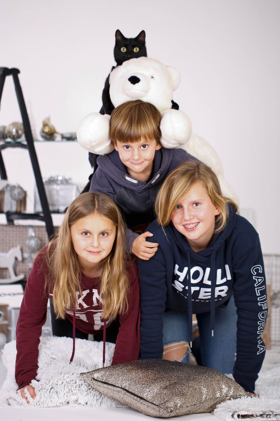 Drei Kinder und eine Katze sitzen auf einem Teddybären und lächeln vor einem Regal.